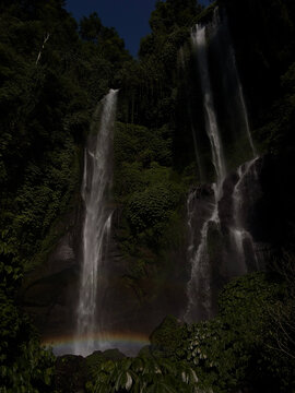 Two majestic waterfalls cascade down rocky cliffs surrounded by dense green foliage. The scene reflects a tranquil natural environment, with mist rising near the base.