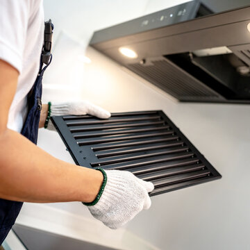 Male hand maintenance worker holding range hood filter in domestic kitchen. Technician in protective gloves removing black metal filter from cooker hood. Home appliance repair service