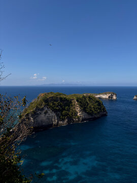 A scenic view of rocky islands in a clear blue ocean. The coastline features lush green vegetation contrasting with the vibrant water. The sky above is bright and sunny.