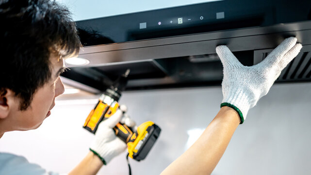 Asian male technician using electric screwdriver fixing extractor motor in cooker hood in modern kitchen. Maintenance worker man repairing exhaust fan in kitchen hood. Home appliance service