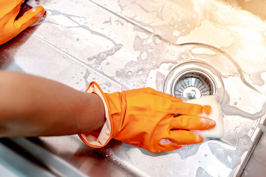 Housekeeper hand wearing orange rubber glove scrubbing on kitchen sink drainer with soap suds spreading around. Cleaning stainless steel sink for hygiene. Housework chores in the kitchen