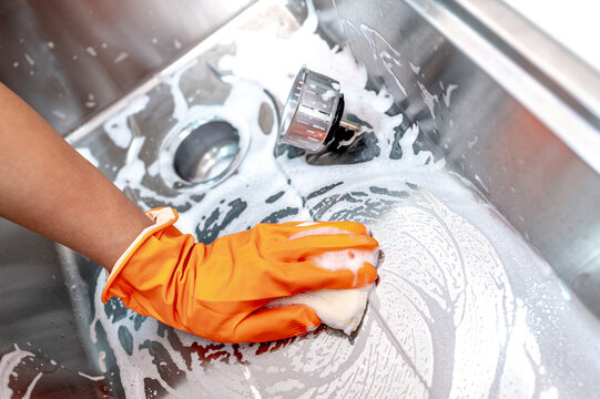 Housekeeper hand wearing orange rubber glove scrubbing on kitchen sink drainer with soap suds spreading around. Cleaning stainless steel sink for hygiene. Housework chores in the kitchen