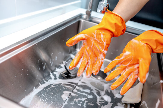 Housekeeper showing hands with orange rubber glove full with soap suds while scrubbing on kitchen sink surface. Cleaning stainless steel sink for hygiene. Housework chores in the kitchen