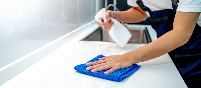 Male housekeeper hand holding detergent spray bottle while wiping blue microfiber cloth on modern white countertop in domestic kitchen. Home hygiene maintenance. Cleaning house service