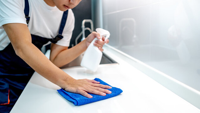 Asian male housekeeper holding detergent spray bottle while wiping blue microfiber cloth on modern white countertop in domestic kitchen. Home hygiene maintenance. Cleaning house service