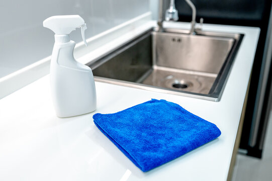 Blue microfiber cloth and detergent spray bottle on modern white countertop near stainless steel sink in domestic kitchen. Cleaning tool product for housework chores and home hygiene