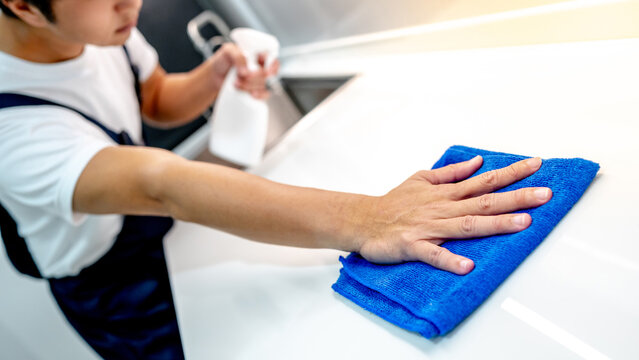 Asian male housekeeper holding detergent spray bottle while wiping blue microfiber cloth on modern white countertop in domestic kitchen. Home hygiene maintenance. Cleaning house service