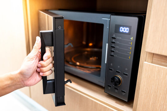 Hand holding door handle opening black microwave oven in modern wooden cabinet. Home appliance in the kitchen