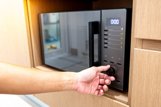 Hand turning knob setting timer on modern black microwave oven in wooden cabinet. Using home appliance in the kitchen