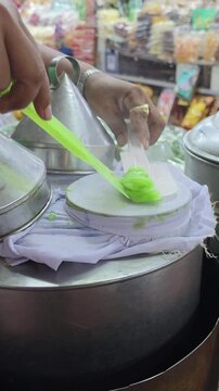 A vertical close-up shot of a local Thai street vendor using a spatula to add sweet peanut and radish filling onto a steamed green rice skin crepe over a traditional cloth pot steamer.