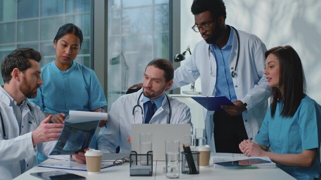 Serious group of multiracial doctors talking at table working together in clinic. Caucasian leader talk advice to interns assistance team coworkers therapists medicine work x-rays papers at hospital