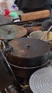 A vertical close-up shot of a street vendor pouring sweet coconut milk batter with black sesame seeds onto a hot iron press to bake traditional Thai crispy coconut roll dessert (Thong Muan Krob).