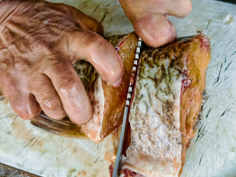 Hands are working to prepare fish on a cutting board in a kitchen. A knife moves carefully along the fish as it is cleaned and cut