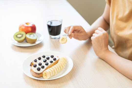 Closeup of Asian woman hand holding fork eating healthy breakfast toast with blueberry, banana, kiwi, red apple and black coffee on wooden table ai=t home, wellness and diet lifestyle concept.