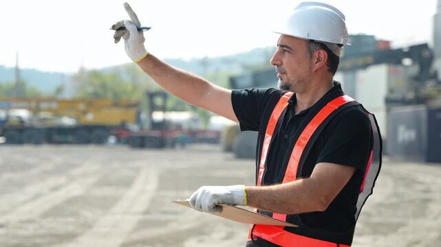 Professional male engineer in a hard hat and safety vest pointing finger while holding a clipboard at a busy construction site or industrial container yard, managing logistics and site operations.