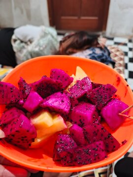 Semangkuk buah naga merah yang sudah dipotong-potong menjadi beberapa bagian dan disajikan dalam mangkuk berwarna oranye. Daging buahnya berwarna merah muda cerah dengan biji-biji hitam kecil yang ter