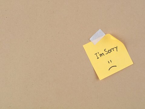 Handwritten note saying I'm sorry with a sad face on a yellow adhesive note taped to a brown cardboard background. Perfect concept for apology, forgiveness, sympathy, and emotional message
