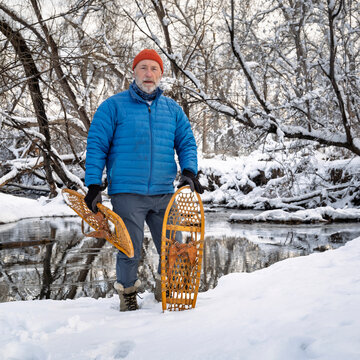 senior male with classic wooden snowshoes (Bear Paw) on a stream shore, winter scenery in Colorado