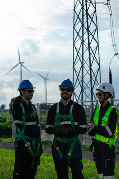 Three wind energy technicians in helmets and safety harnesses coordinate beside an electrical substation, with wind turbines behind them, representing teamwork, grid maintenance and renewable power.