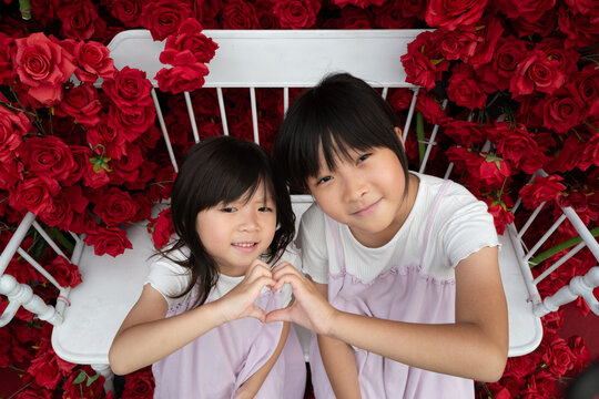 Two Girls Make a Heart Shape With Their Hands Among Red Roses