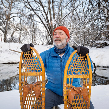 senior male with classic wooden snowshoes (Bear Paw) on a stream shore, winter scenery in Colorado