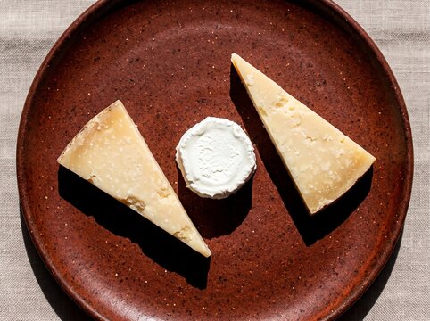 Minimalist presentation of hard aged cheese wedges and soft goat cheese round on textured brown ceramic plate for modern food photography and culinary branding.