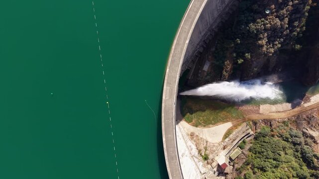 Overhead view of the curved crest of Baells Dam in Catalonia, Spain, releasing a powerful jet of water into a rocky gorge, with turquoise reservoir waters alongside