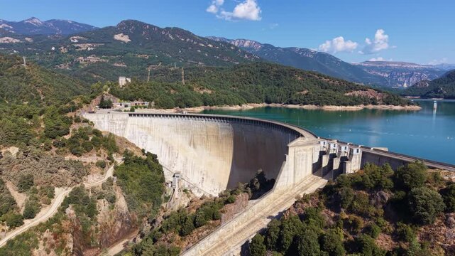 Aerial view of Baells Dam curving across a rocky gorge, holding a turquoise reservoir in Catalonia, Spain, with forested mountains, cliff bands, and a ruined hilltop castle in the distance