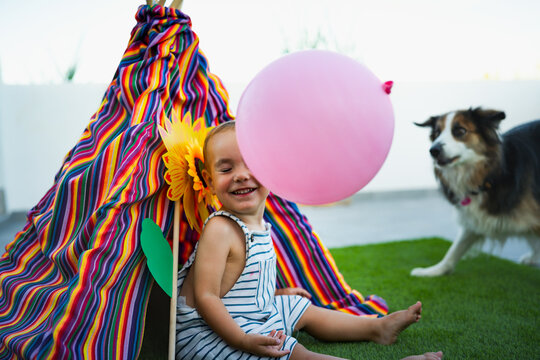 Toddler smiling, playing with balloon, dog watching outside