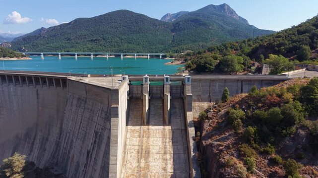 Aerial view of concrete spillway gate structures at Baells Dam in Catalonia, Spain, overlooking a turquoise reservoir, road bridge, and lush forested mountains under blue sky