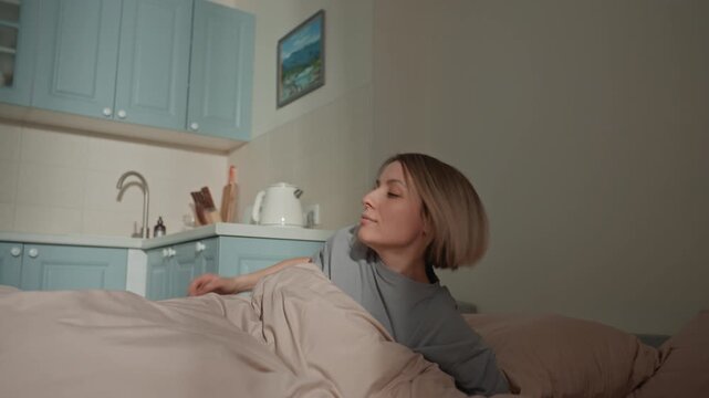 Woman waking in pastel kitchen bed. Intimate morning sequence showing bedroom to kitchenette transition short blonde woman sits up, stretches, walks toward blue cabinets, opens cupboards, checks