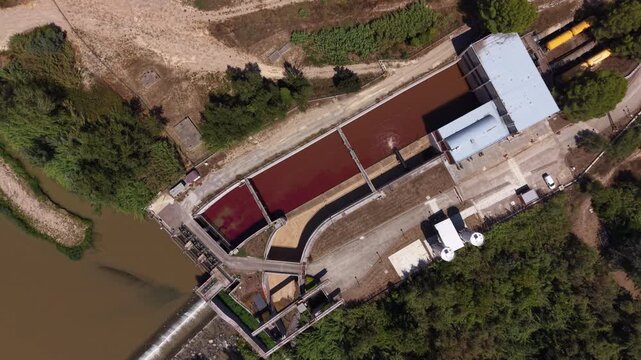 Top view of a drinking water treatment plant intake channel showing deep red-burgundy water dosed with potassium permanganate for pre-oxidation, flowing from the adjacent river into treatment basins