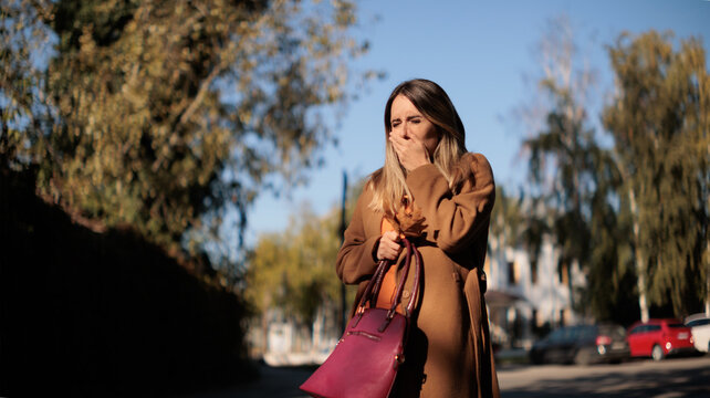 Sick Young Woman Outdoors Sneezing Or Coughing Covering Her Mouth With Hand. Unwell female model squinting in discomfort while catching cold symptoms on autumn city street