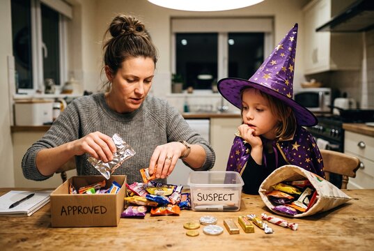 A woman sits at a table sorting through piles of candy while a child watches. They are preparing Halloween treats in a cozy kitchen setting during the night