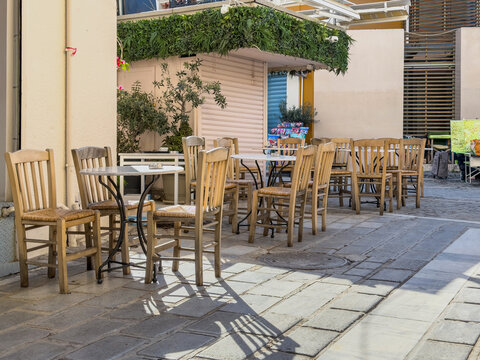 Empty wooden outdoor cafe seating beneath leafy greenery on pedestrian lane in Monastiraki, Athens