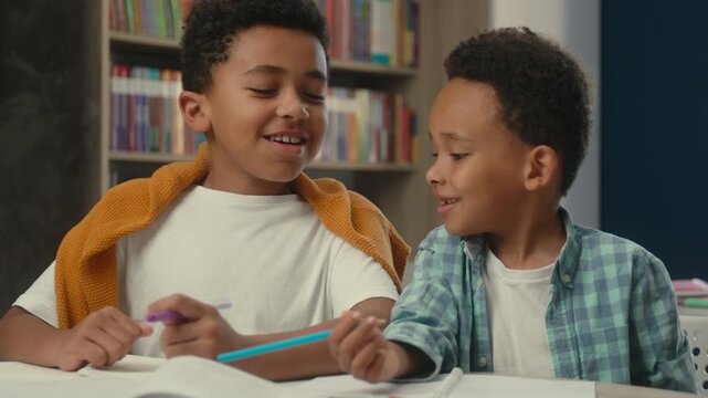 Two African American happy kids little boys writing homework write at lesson at school elementary education class talking siblings brothers pupils sons studying learning together talk study learn