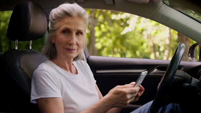 Serious Caucasian old woman using mobile phone navigator inside car looking at camera retired female senior lady chatting smartphone look with confidence sitting in luxury own auto rent automobile
