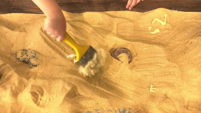 Child using a brush to clear sand while searching for hidden objects at an interactive exhibit, educational play, hands on learning.