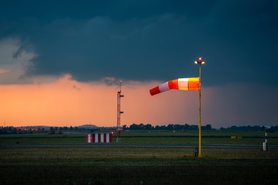 Red and white striped windsock fully extended by strong wind at airport airfield against oncoming dark storm clouds. Severe weather warning, aviation safety and meteorology forecast concept.