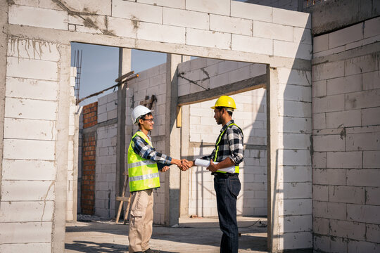 Asian Civil Engineers Handshake on Construction Site with Blueprint and Wearing Safety Uniform and Hard Hats at Building Structure for Project Progress