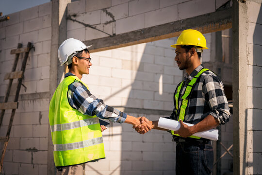 Two Adult Construction Workers Shaking Hands at Building Site with Hard Hats Safety Vests and Blueprint Plans Construction Partnership Concept