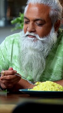 Indian mid age man watching smartphone reels while eating at home, stylish bearded male enjoying poha at breakfast table and streaming web series or comedy videos in modern kitchen lifestyle