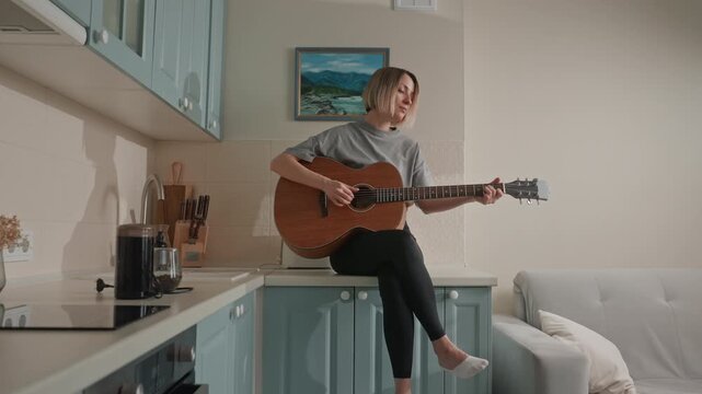 White woman playing guitar in kitchen, soft acoustic practice on blue cabinets counter, barefoot, calm singersongwriter rehearsing, intimate home atmosphere with painting and sofa background, music