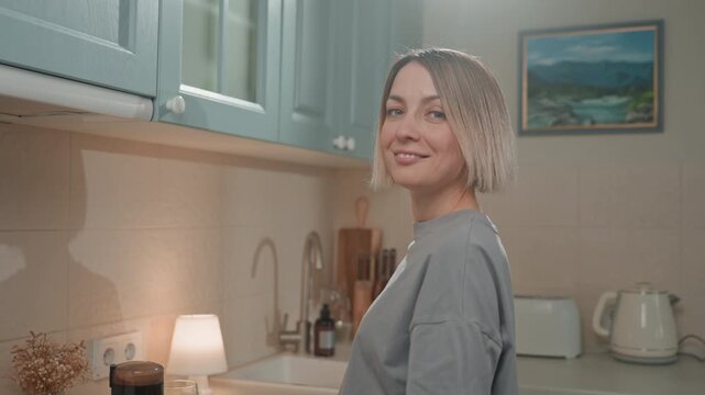 White woman making coffee in bright kitchen, pouring espresso at counter. Morning sunlight fills pastel cabinets, toaster and kettle visible on shelf. She smiles and hums a tune, joyful music lover