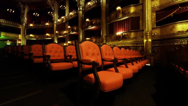 Dimly lit rows of empty seats with amber glow, plush upholstery and deep shadows, ornate boxes overhead, aisle markers illuminated, quiet theatrical atmosphere