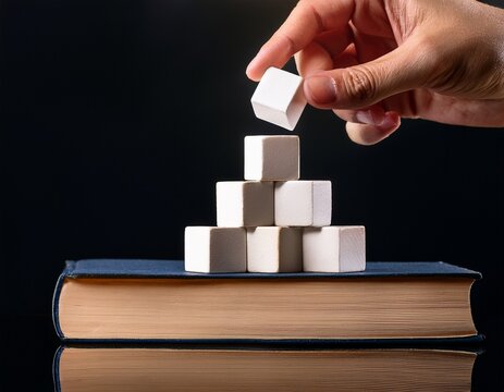 hand placing a white cube on a pyramid stack on a book