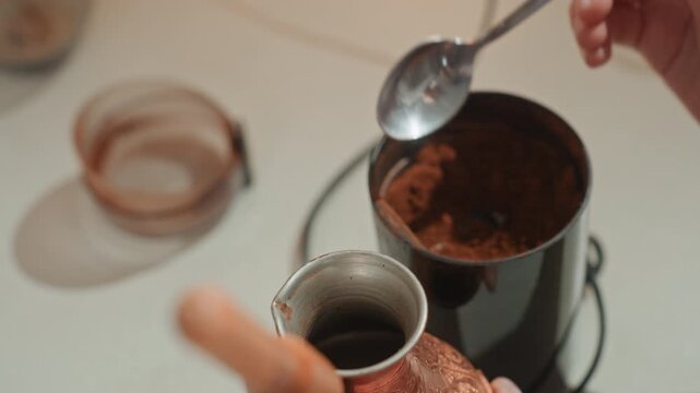 Spoon scooping coffee grounds from canister, white hands transferring measured scoop to copper cezve on counter, black container and lid visible, precise kitchen routine and rhythmic motion, focused
