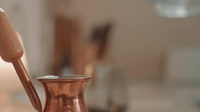 Closeup copper cezve receiving gentle pour, measured stream and soft reflections on metal, tabletop composition with blurred background, apartment barista and craft enthusiast creating ASMR rhythm
