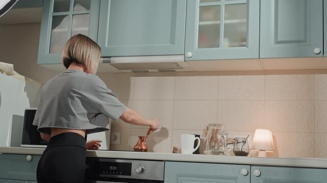 Caucasian woman in kitchen preparing coffee, music lover mood and slow domestic ritual at stove, stirring brew with steady hands, warm lamp glow, pastel cabinets and cozy shadows create relaxed