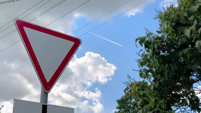  Triangular yield road sign with red border against blue sky with clouds and green tree, give way sign, traffic warning, road safety, urban transport.
